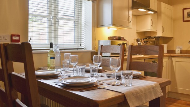 A dining table set with plates, glasses, and cutlery in a room with wooden chairs, a window with blinds, and a kitchen area with cabinets and appliances. - 2 bedroom accommodation in Myddle, near Shrewsbury (Myddle, near Shrewsbury)