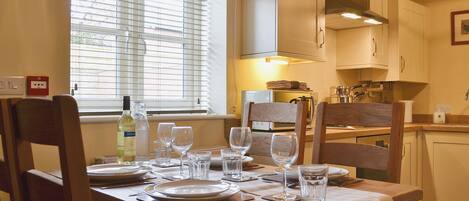 Una mesa de comedor dispuesta con platos, vasos y cubiertos en una habitación con sillas de madera, una ventana con persianas y una zona de cocina con armarios y electrodomésticos.