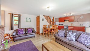 A living room with a brown leather sofa, a wooden coffee table, and a purple rug. There is a dining area with a wooden table and chairs. The kitchen has red cabinets and white appliances. - 2 bedroom accommodation in Alston (Alston)