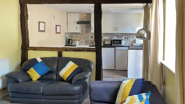 A living room with a sofa and armchair, a kitchen in the background, and a window with curtains. - Orchard Bank Cottage (Little London, near Gloucester)