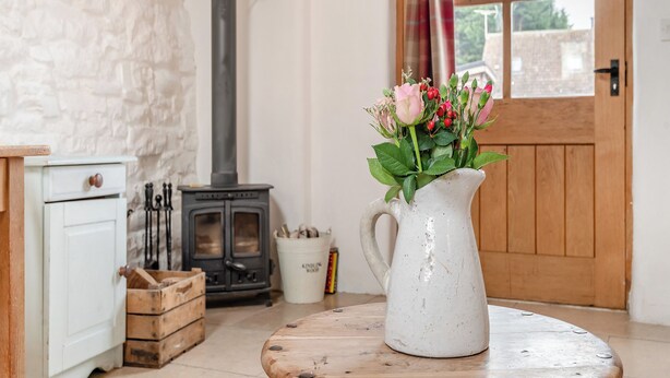 A rustic room with a white stone wall, a wood-burning stove, a wooden door, and a white pitcher with flowers on a round wooden table.