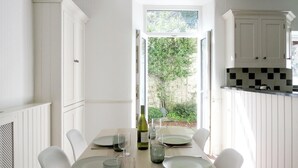 Een eetgedeelte met een tafel voor vier personen, witte stoelen, een raam met uitzicht op groen, en een keuken met witte kasten en een zwart-wit geruit spatwand.