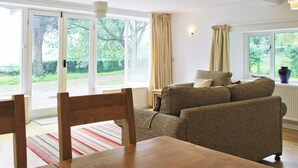 Un salon avec un canapé, une table en bois et des chaises. De grandes fenêtres, ornées de rideaux, offrent une vue sur la verdure environnante.