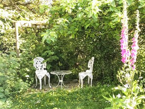 A garden with white metal chairs and a table, surrounded by greenery and flowering plants.