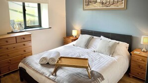 A neatly made bed with a wooden tray, a painting of a forest on the wall, and a window with blinds. - Red Kite Cottage (Heyope, near Knighton)