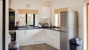 A modern kitchen with white cabinets, a stainless steel refrigerator, a microwave, and a sink. There are two trash bins and a window with a patterned valance. - Red Kite Cottage (Heyope, near Knighton)