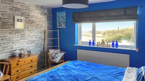 A bedroom with a blue bedspread, a wooden dresser, a chair with an anchor pillow, a window with bottles, and a view of the outdoors.