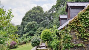 Un bâtiment en pierre doté d’un toit en pente, entouré d’un jardin luxuriant où se trouve un banc en bois.