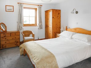 A bedroom with a wooden bed, a wooden dresser, a chair, a wardrobe, a window with curtains, and a wall-mounted lamp.