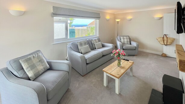 A living room with a grey sofa, a smaller armchair, a wooden coffee table with a vase of flowers, and a large window with a view of the outside. - 2 bedroom accommodation in Port Isaac (Port Isaac)