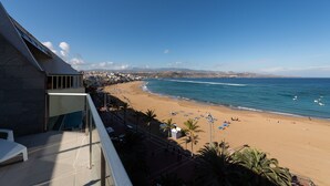 Vista de la playa desde un balcón, cielo despejado, arena y algunas embarcaciones en el agua.