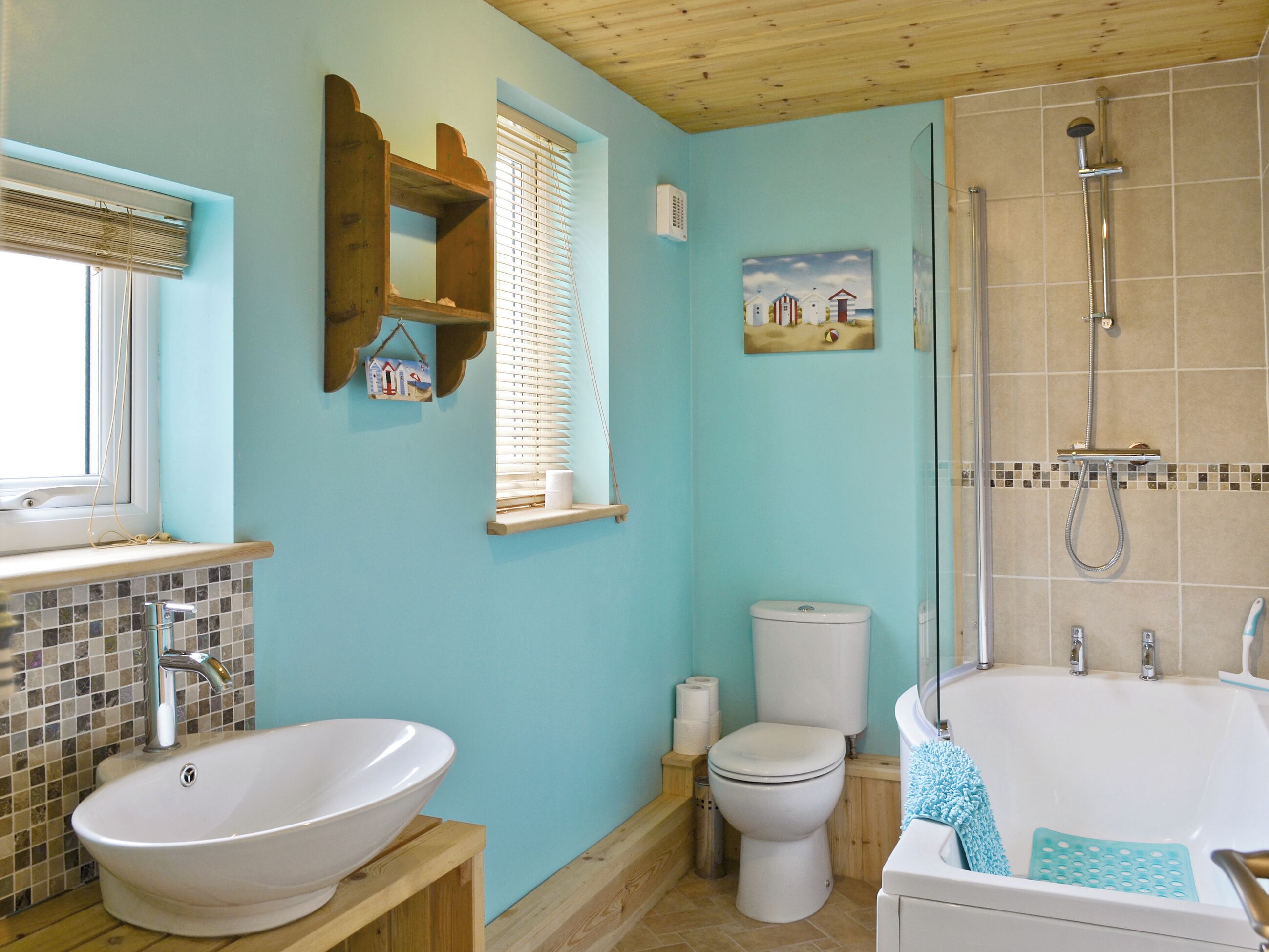 Een badkamer met een witte wastafel, toilet en badkuip. Er is een raam met jaloezieën, een houten plank en een schilderij aan de muur.
