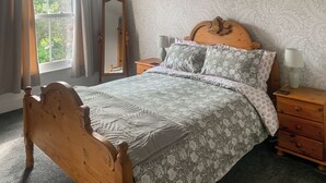 Une chambre à coucher avec un lit en bois, une literie à motifs floraux, un miroir, une table de chevet avec une lampe et une fenêtre avec des rideaux.