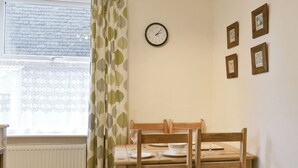 Un coin repas avec une table en bois, des chaises, une horloge et une fenêtre ornée d’un rideau à motifs de feuilles.