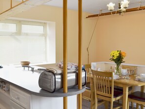 Een keuken met een broodrooster, een eettafel met stoelen, een vaas met bloemen en een raam met jaloezieën.