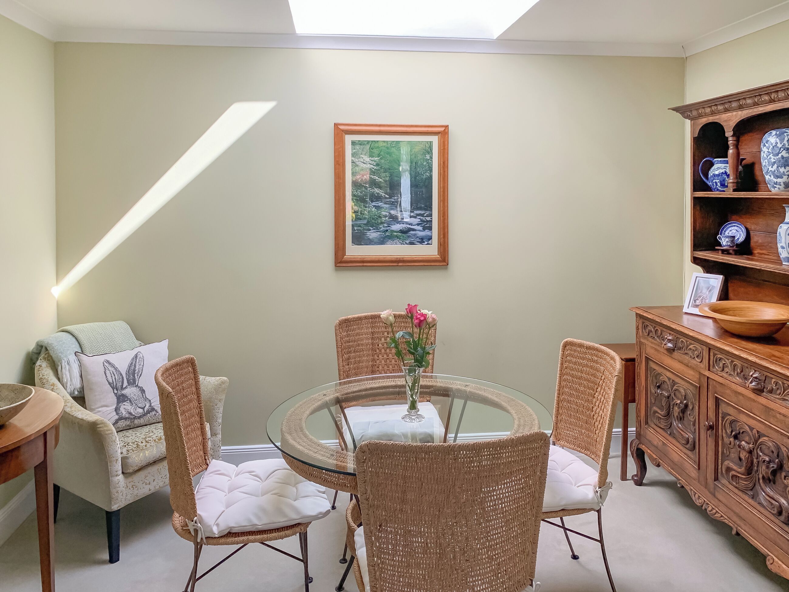 A dining area with wicker chairs, a glass table, a vase with flowers, a wooden cabinet with dishes, and a framed picture on the wall.