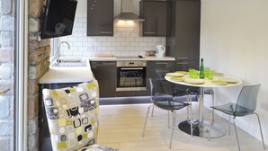 A compact kitchen with a dining area, a flat-screen TV mounted on the wall, and a stone wall on one side. - 2 bedroom accommodation in Lelant, near St Ives (Lelant, near St Ives)