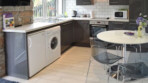 A modern kitchen with a built-in washing machine, a dining table with chairs, a TV mounted on the wall, and a window with a view of the outside. - 2 bedroom accommodation in Lelant, near St Ives (Lelant, near St Ives)