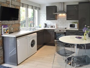 A modern kitchen with a built-in washing machine, a dining table with chairs, a TV mounted on the wall, and a window with a view of the outside. - 2 bedroom accommodation in Lelant, near St Ives (Lelant, near St Ives)