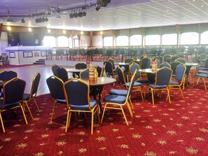 Une salle de banquet avec des tables et des chaises rondes, une scène et de grandes fenêtres.
