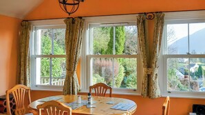 Un coin repas avec une table ronde en bois et quatre chaises, de grandes fenêtres avec des rideaux et une vue sur la verdure à l’extérieur.