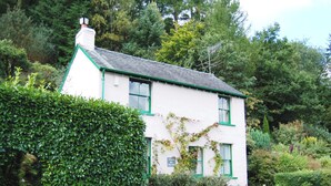 Une petite maison blanche aux boiseries vertes, avec des fondations en pierres apparentes, entourée d’une végétation luxuriante et d’arbres.