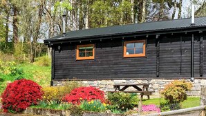 Una cabaña de madera con dos ventanas, zócalo de piedra y una mesa de picnic al frente.