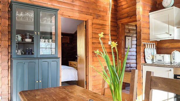 Interior de una cabaña de madera con una mesa de comedor, un aparador con puertas de vidrio, una zona de cocina y una cama al fondo.