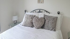 Ein ordentlich bezogenes Bett mit einer gestreiften Tagesdecke und dekorativen Kissen, darunter ein herzförmiges, in einem schlicht eingerichteten Zimmer.