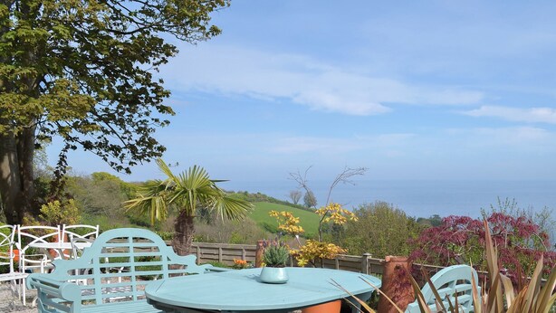 Eine Terrasse mit rundem Tisch und zwei Stühlen, umgeben von Pflanzen und Blick aufs Meer.