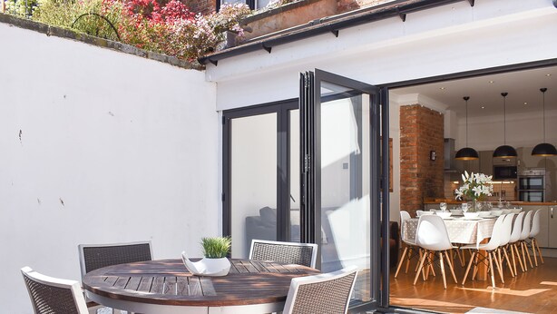 Un patio al aire libre con una mesa redonda de madera y sillas de mimbre blanco, una planta pequeña en maceta y un ventanal de vidrio que da a un comedor con mantel blanco y sillas.