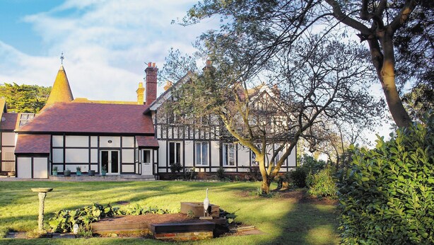 A traditional Tudor-style house with a steeply pitched roof, a prominent chimney, and a well-maintained garden with a stone birdbath and a wooden bench. - Bodwen Corner Cottage (Wootton Bridge)