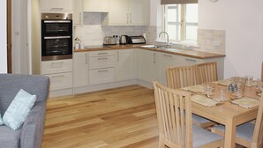 A modern kitchen with white cabinets, a built-in oven, and a dining area with wooden chairs and a table set for four. - Bodwen Corner Cottage (Wootton Bridge)