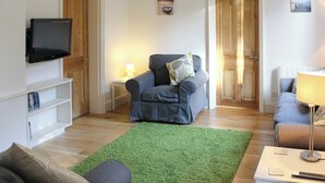 A living room with a flat-screen TV, a grey sofa, a green rug, and a wooden floor. - Bodwen Corner Cottage (Wootton Bridge)