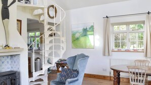 Een knus woongedeelte met een open haard, een blauwe fauteuil, een houten eettafel en een wenteltrap.