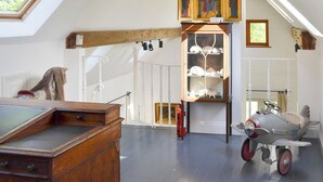 Een kamer met een schuine plafond, een houten bureau, een plank met decoratieve objecten, een klein speelautootje en een wandplank met kristallen glazen.