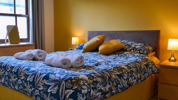 A hotel room with a large bed, floral bedding, two pillows, a bedside lamp, and a window with curtains. - Bleaberry- Hewetson Court (Keswick)