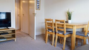 A room with a wooden dining table, four chairs, a television on a stand, and a door leading to another room. - Bleaberry- Hewetson Court (Keswick)
