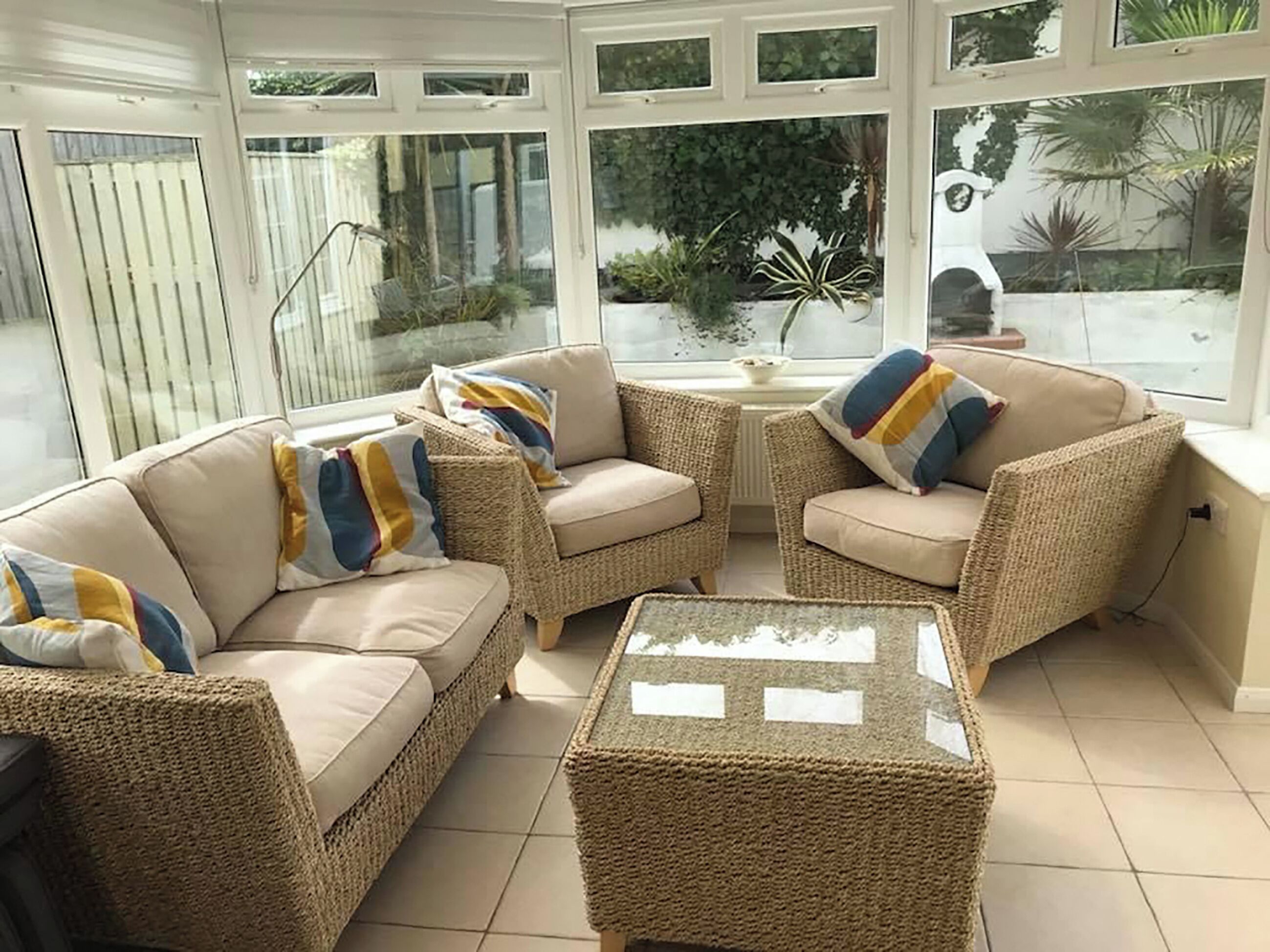 A sunlit room with wicker furniture, large windows, and a view of greenery outside.
