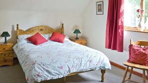 Une chambre à coucher avec un lit en bois, une literie à motifs floraux, deux tables de chevet avec des lampes, une chaise, une fenêtre avec des rideaux rouges et un tableau encadré accroché au mur.