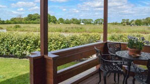 A covered outdoor seating area with a wooden bench and metal chairs, overlooking a grassy field and a body of water. - 3 bedroom accommodation in Burgh le Marsh, near Skegness (Burgh le Marsh, near Skegness)