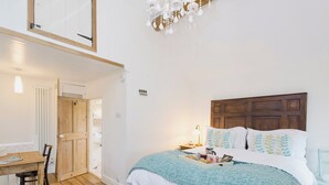 Une chambre avec un plancher en bois, un lit recouvert d’une couverture blanche et bleue, une tête de lit en bois, une table de chevet avec une lampe et un bureau en bois avec une chaise.