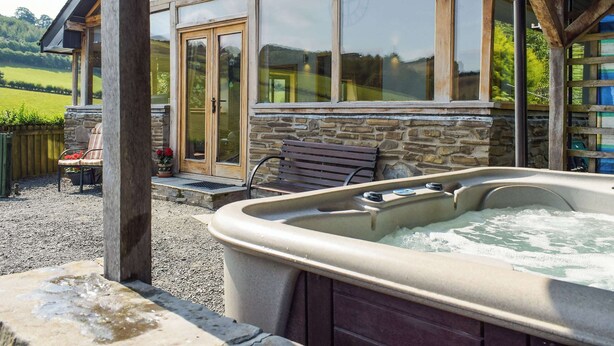 A spa area with a hot tub, stone walls, wooden beams, and a view of a green landscape.