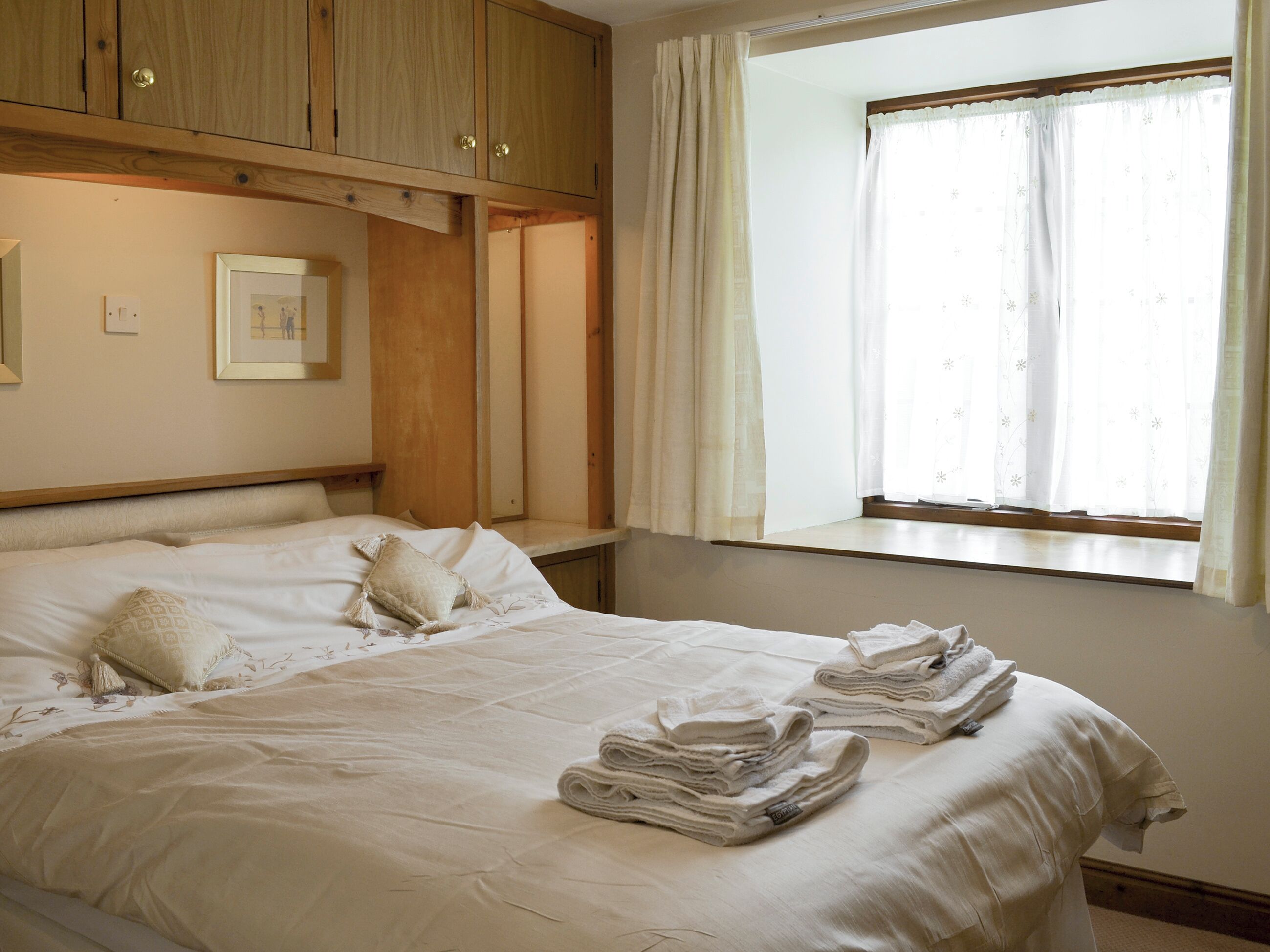 A neatly made bed with pillows and folded towels, wooden headboard, and a window with curtains.