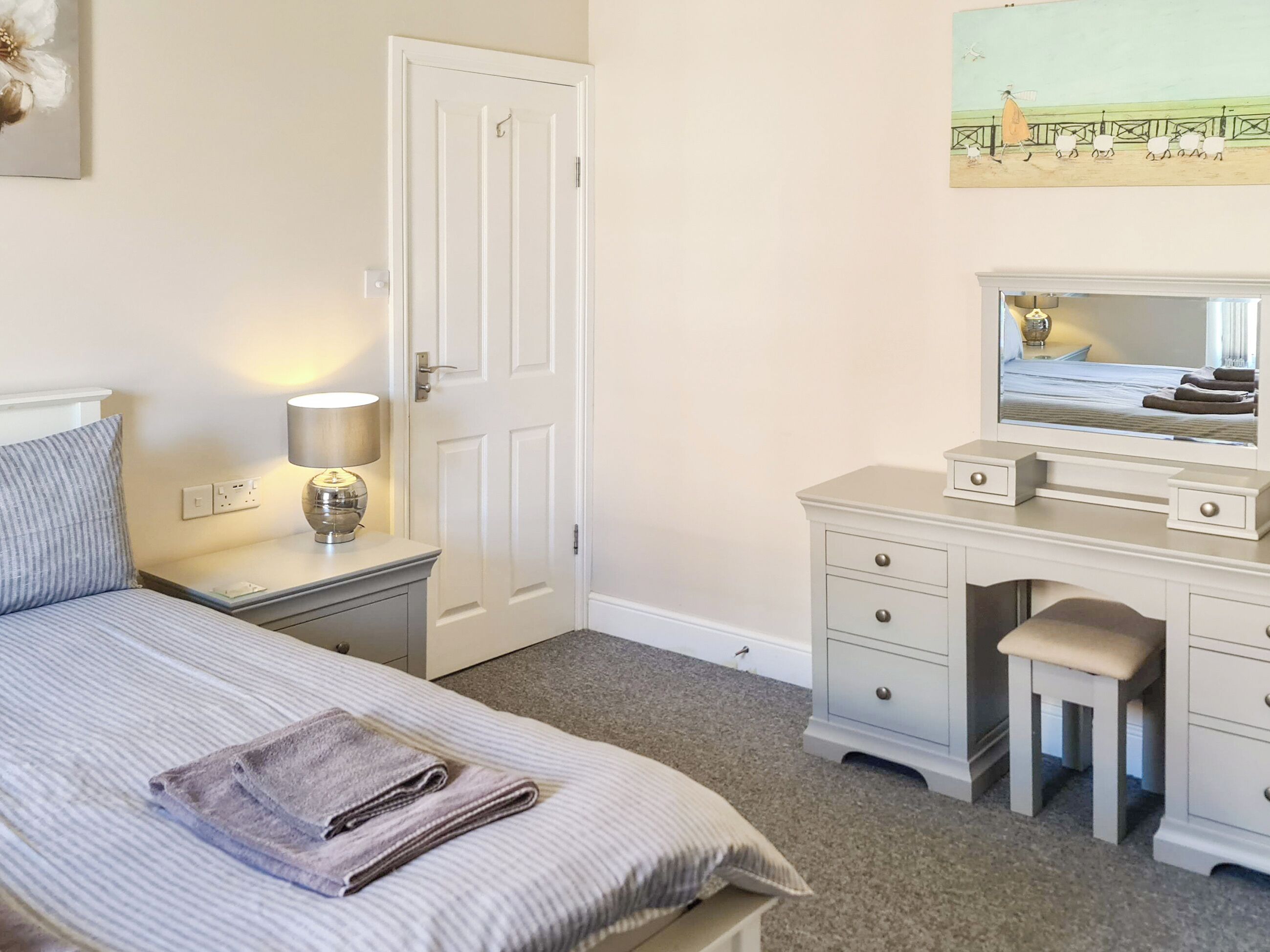 A bedroom with a bed, bedside table, lamp, dresser, mirror, and a painting on the wall.
