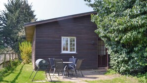 A wooden cabin with a patio area, a grill, and a table with chairs. - 2 bedroom accommodation in Scarning, near Dereham (Scarning, near Dereham)