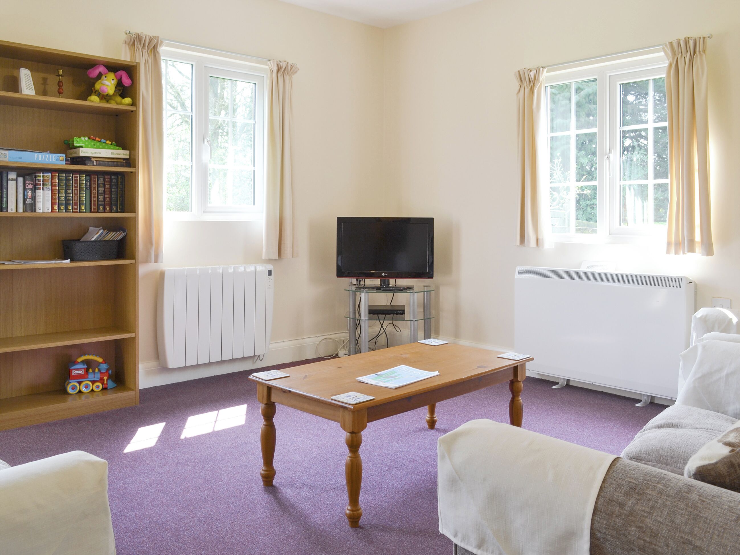 A living room with a wooden bookshelf, a television on a stand, a table with papers, a radiator, and a window with curtains.