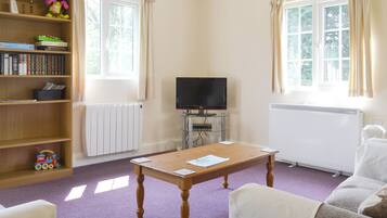 A living room with a wooden bookshelf, a television on a stand, a table with papers, a radiator, and a window with curtains.