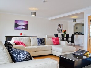 Un salon moderne avec un canapé d’angle, un coin repas avec des chaises, une cuisine visible à travers une arche et un tableau accroché au mur.