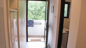 En hall som leder ut till en balkong med utsikt över träd, en dörr med ett handtag och en liten ventilationsöppning i väggen.